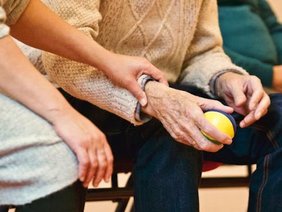 Detailansicht. Zwei Hände einer jungen Frau stützen die Hand einer älteren Person, die einen Ball in der hält.