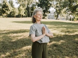 Ältere Frau steht in der Natur und macht eine Bewegung aus dem Yoga. Sie presst die Hände zusammen und hat die Augen geschlossen.