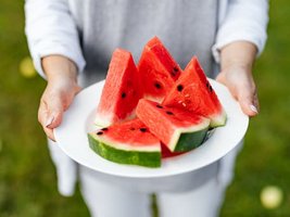 Zwei Hände halten einen Teller mit Wassermelone im Garten. 