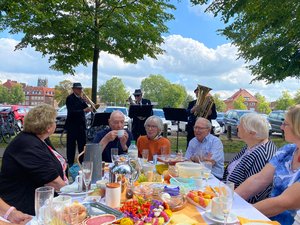 Mitglieder des Vereins sitzen an einem gedeckten Frühstückstisch im Freien. Drei Musiker spielen im Hintergrund ein Lied.